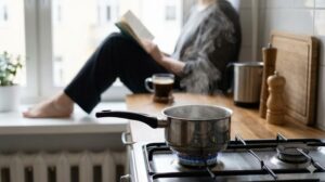 コンロで鍋が煮える横で窓辺で読書しながらコーヒーを飲む女性