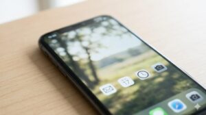 木製デスクの上に置かれたスマートフォンの画面が点灯し、静かな風景壁紙とシンプルなアプリアイコンを表示している。壁紙がアイコンの土台となり、画面全体のまとまりを作り出している様子を示すリアルな写真。